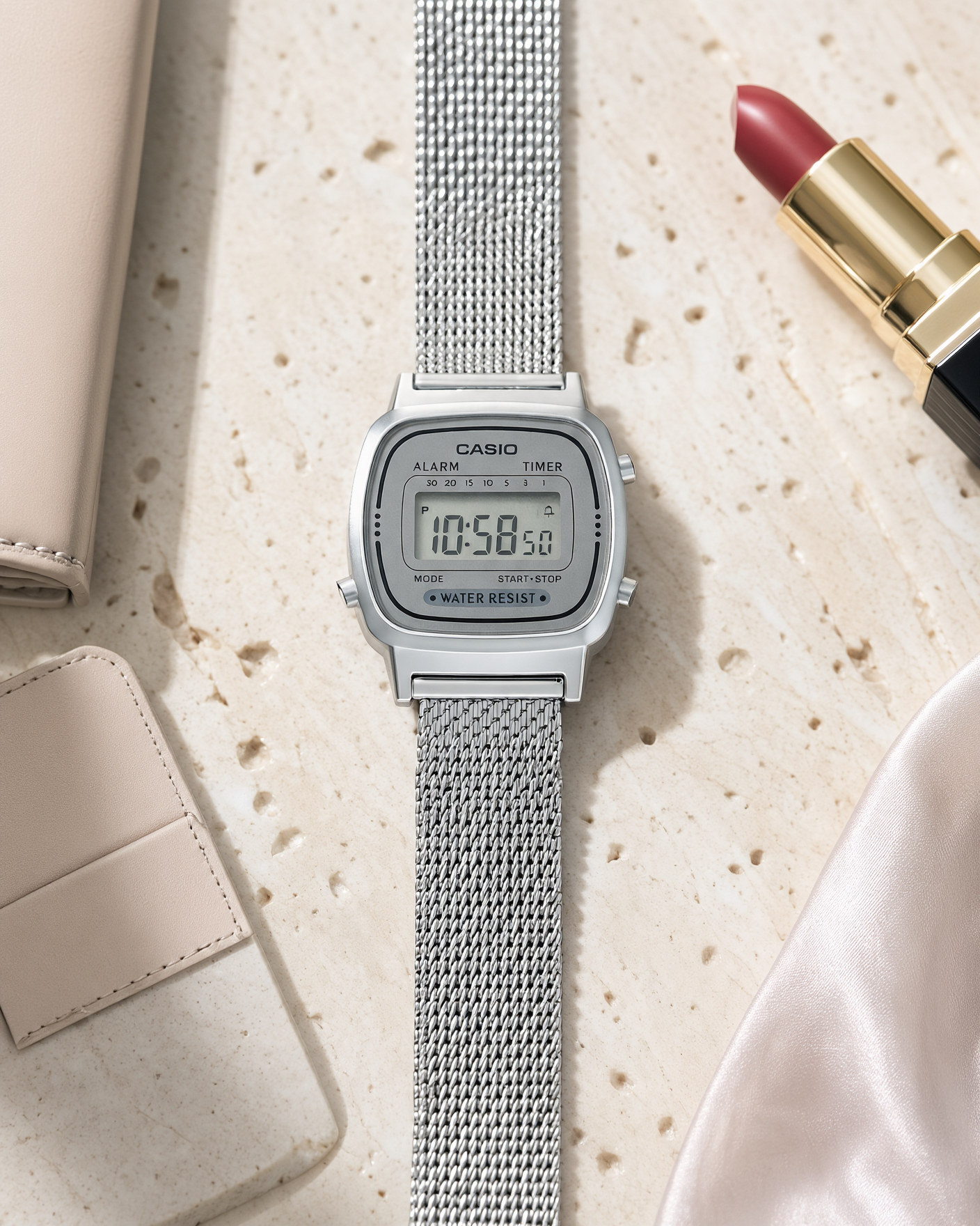 Casio Vintage LA670WEM-7EF women's digital watch with a silver Milanese mesh strap, lying on a travertine tray next to a lipstick and a leather card holder