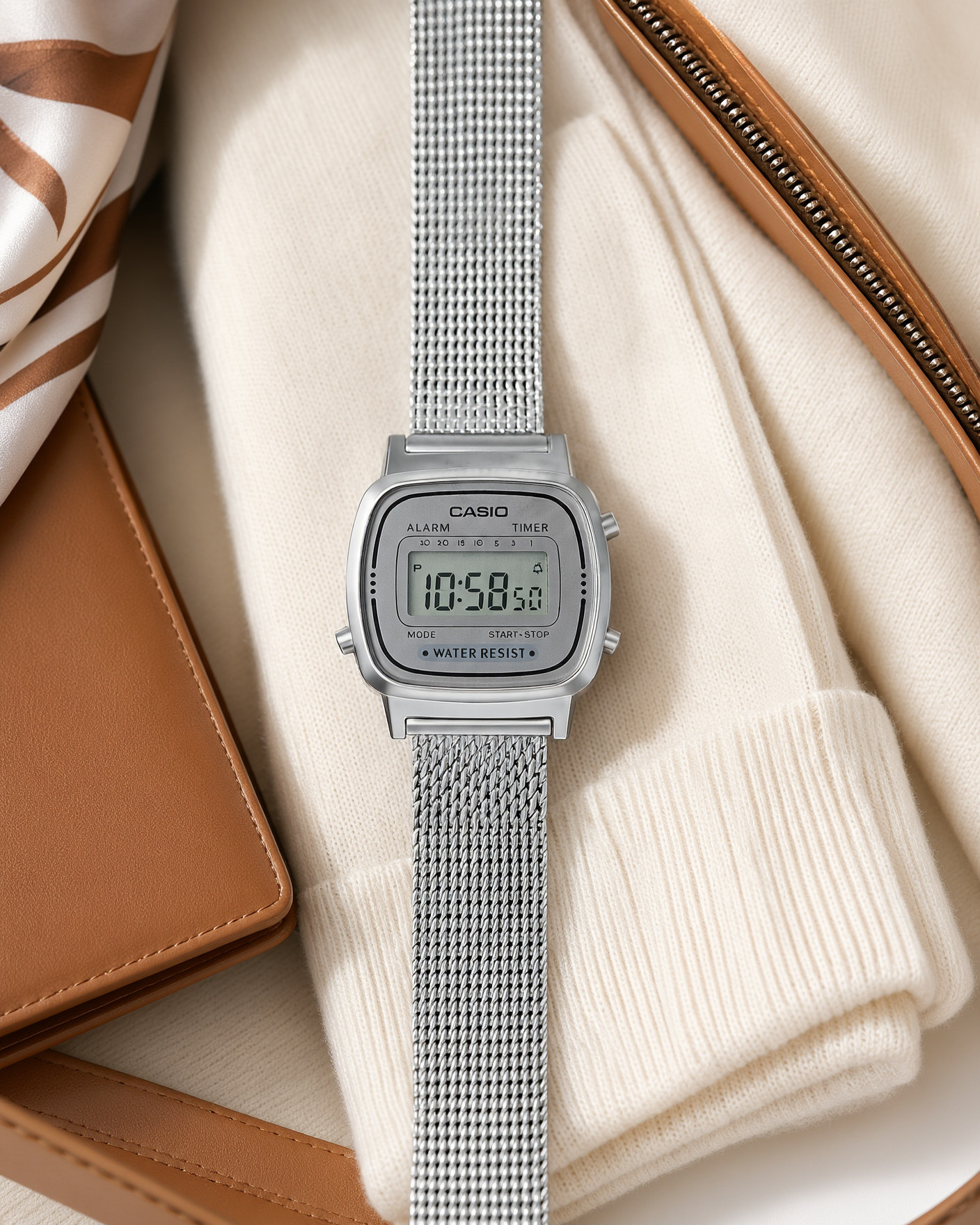 Casio Vintage LA670WEM-7EF silver Milanese mesh watch lying on a cream cashmere sweater inside a leather travel bag, next to a silk scarf