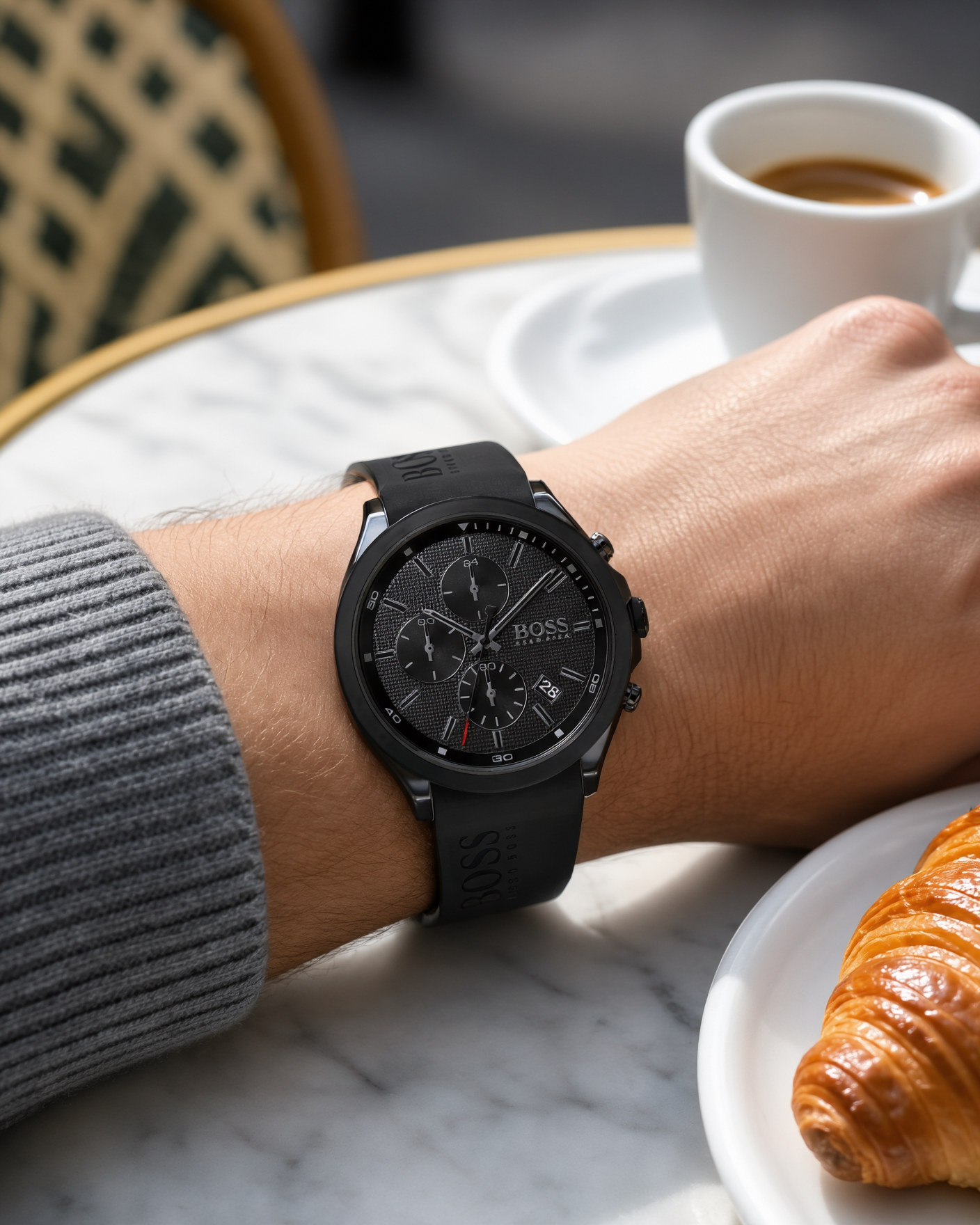 Black Hugo Boss Velocity 1513720 chronograph watch with a silicone strap worn on a man's wrist, on a marble café table with an espresso and a croissant