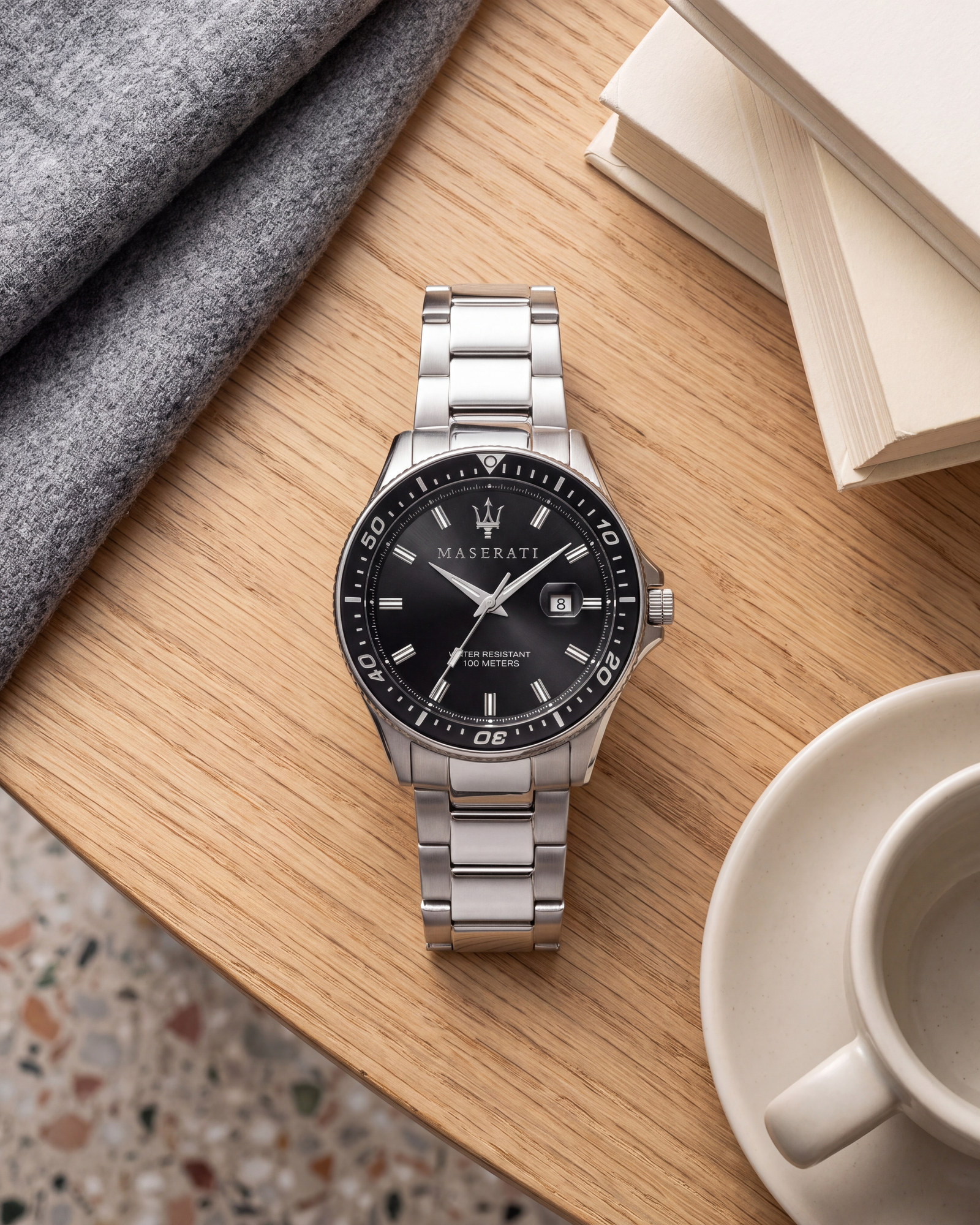 Maserati Sfida R8853140002 watch with black dial and silver stainless steel bracelet, lying on a wooden table beside books and a coffee cup.