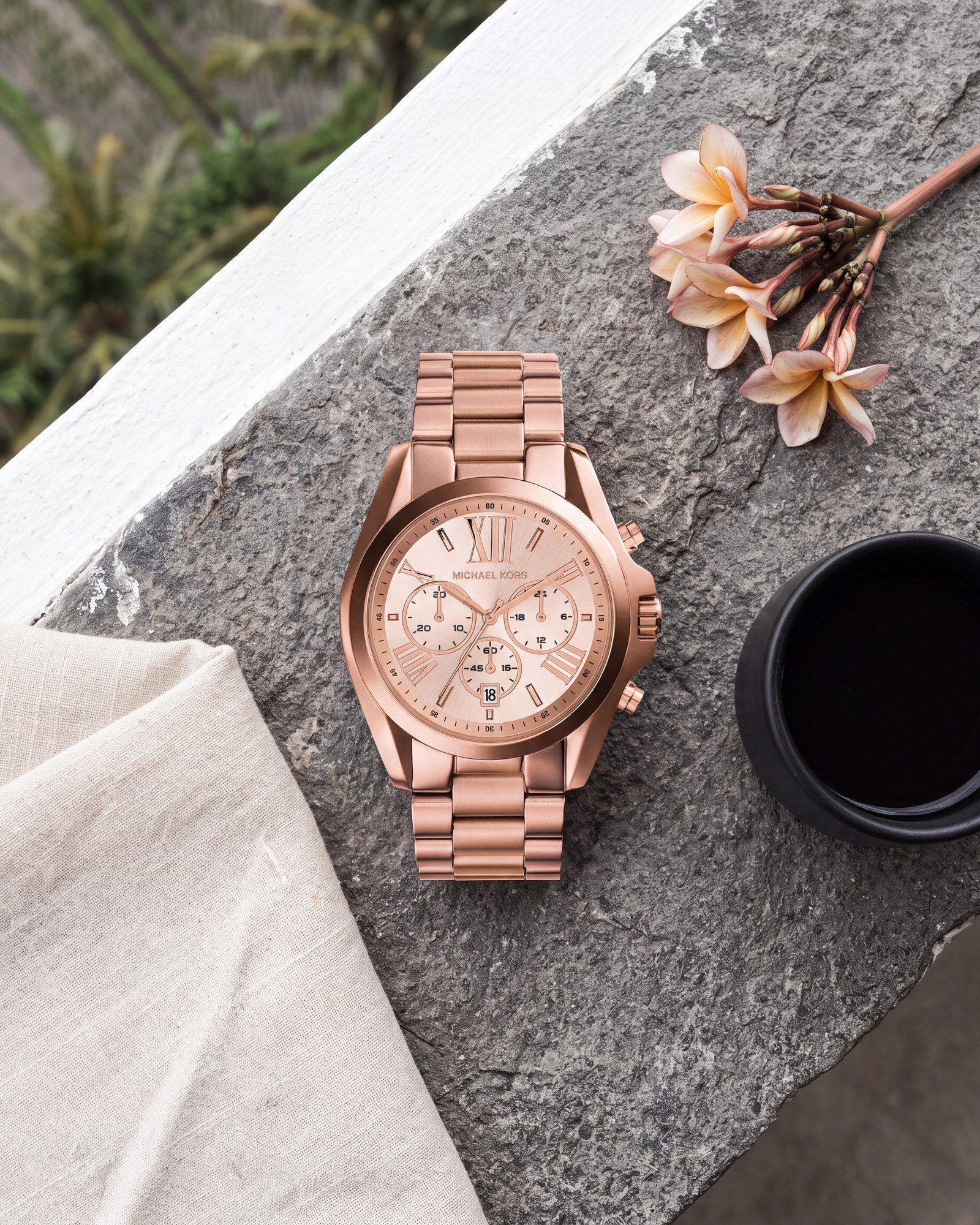 Montre Michael Kors Bradshaw MK5503 posée sur une pierre grise, cadran or rose, bracelet acier or rose, à côté de fleurs de frangipanier et d'une tasse noire.
